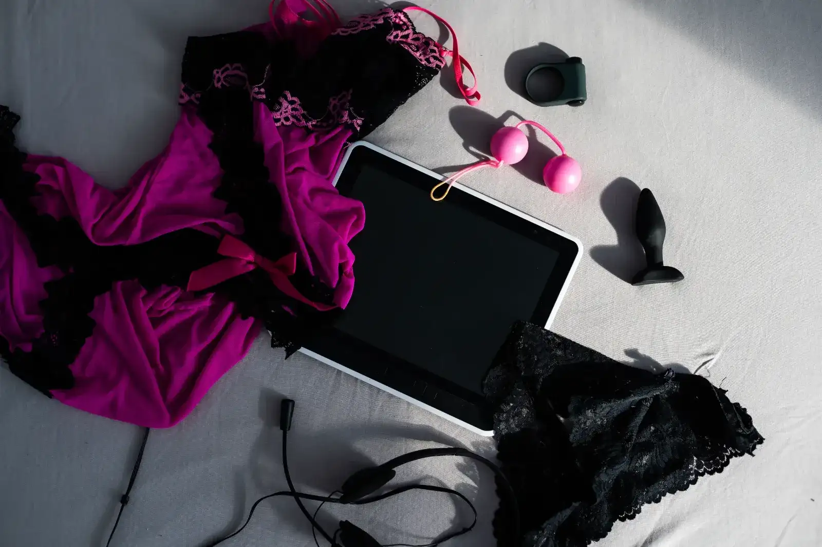 sex shop en ligne, achat et livraison discrète : une tablette posée au milieu d'un lit au draps blancs, une collection de sextoys noirs et roses et de lingeries sexy noires et violettes